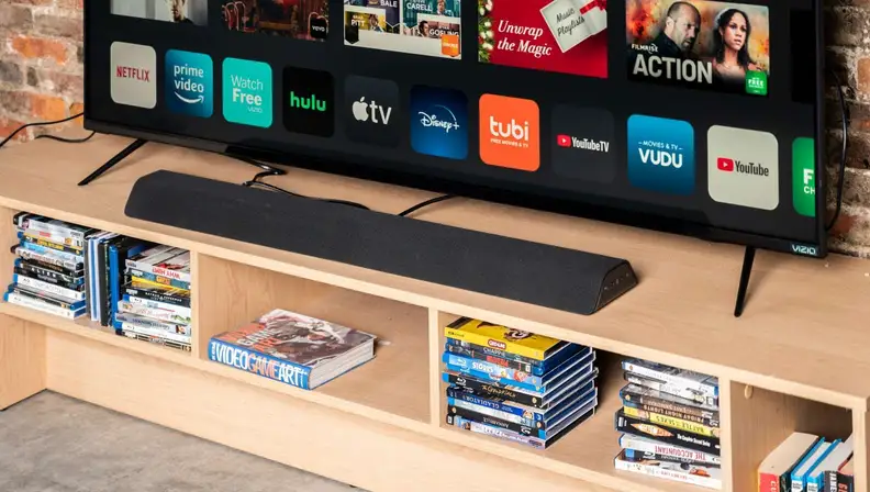 A long trapezoidal soundbar sits between a television and a DVD collection.