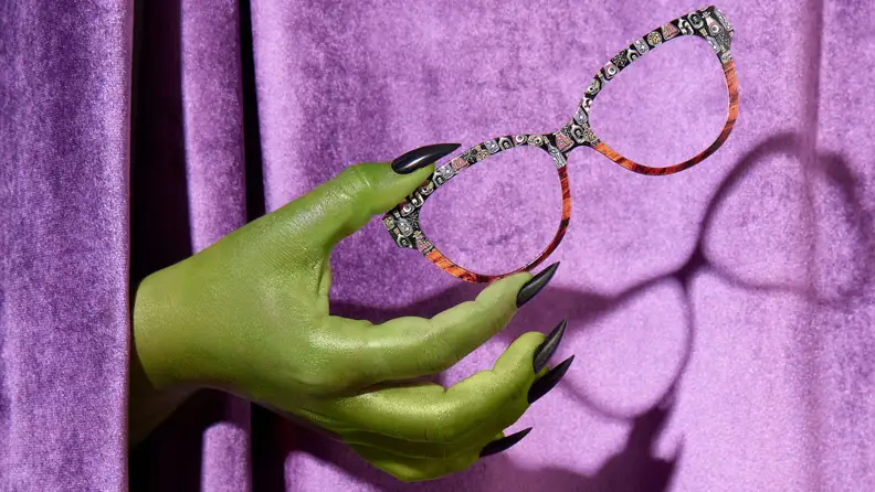 Witches hand holding Halloween-themed eyewear on purple background.