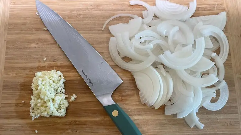 Large knife sitting in between pile of sliced onions and minced garlic on top of wooden cutting board.