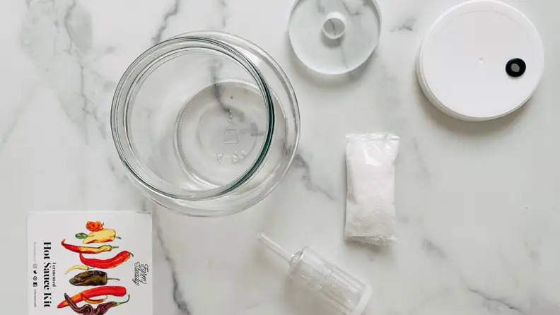 A container and other tools for making fermented hot sauce sit on a marble surface.