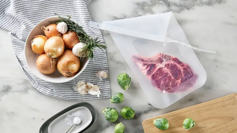 A piece of steak is inside a Stasher silicone bag surrounded by a bowl of onions, garlic, herbs, Brussels sprouts.