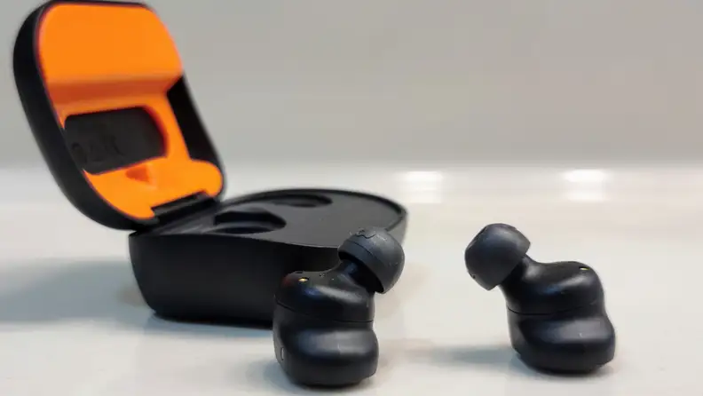 The all-black Skullcandy Grind Fuel sit in front of their orange-lined case on a white reflective glass table with the eartips pointed up.