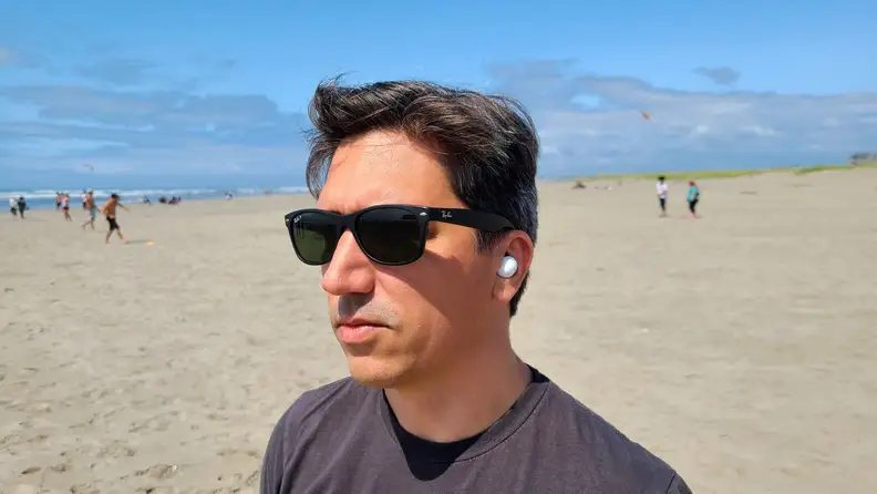 Samsung Galaxy Buds 2 is shown in the ear against a sunny beach, the listener is a man with brown hair wearing sunglasses.