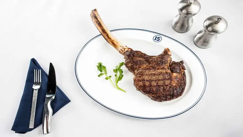 Long bone ribeye with garnishing on a plate, salt and pepper shakers, and fork and knife on a blue cloth napkin