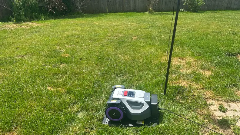 Anthbot M9 robot lawn mower shown sitting on green grass in front of a wooden fence.