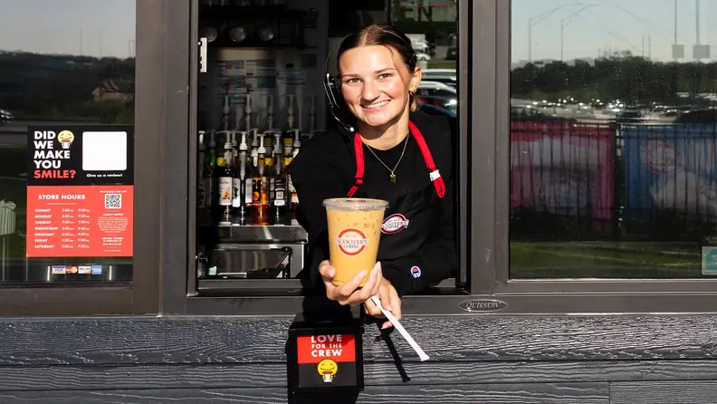 Smiling barista handing an iced coffee through a Scooter's Coffee drive thru window