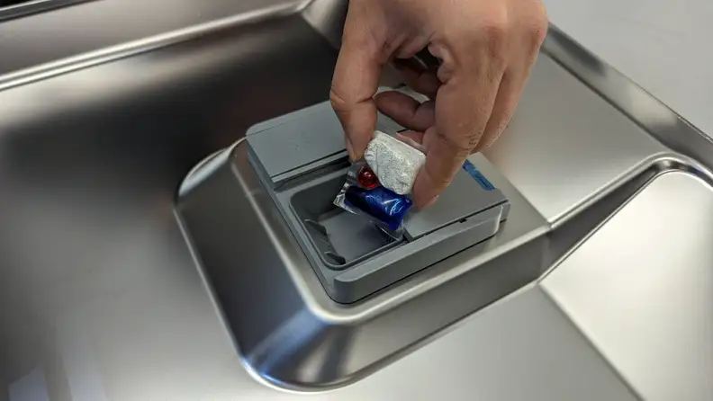 A multicolored dishwasher tablet being placed into a dishwasher detergent dispenser.