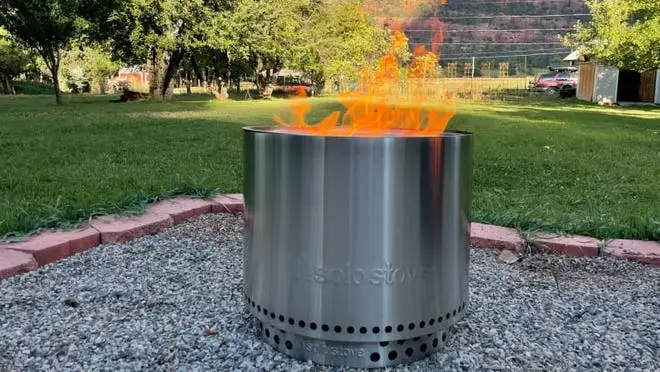 Silver flaming fire pit outdoors on gravel surface.