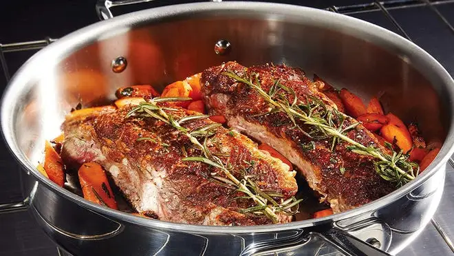 An All-Clad pan on a stove containing ribs on a bed of carrots and topped with rosemary.