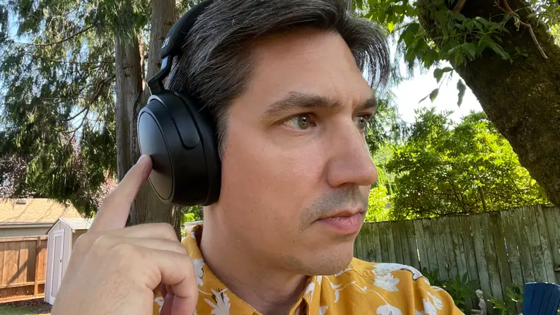 A man in a yellow pattern shirt uses the touch controls on a black pair of headphones in a green yard.