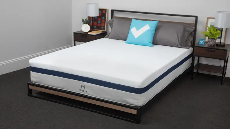 Helix Midnight Mattress without sheets inside of bed frame with three pillows on top, in between two nightstands with lamps and in front of white wall.