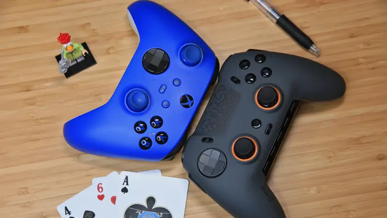 A Blue Xbox gaming controller next to a dark gray Scuf Envision Pro gaming controller, three gaming cards, a pen and a small action figure all on top of wooden surface.
