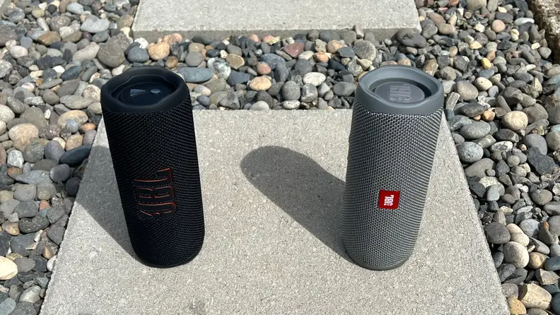 A black, cylindrical Bluetooth speaker sits next to a grey Flip 5 on a concrete paver in gravel.