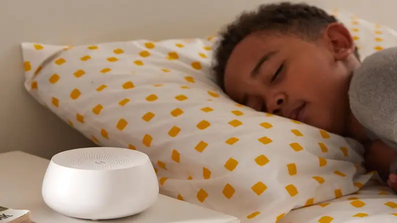 A child sleeps in bed next to a Hatch Rest Mini, a circular white sound machine.