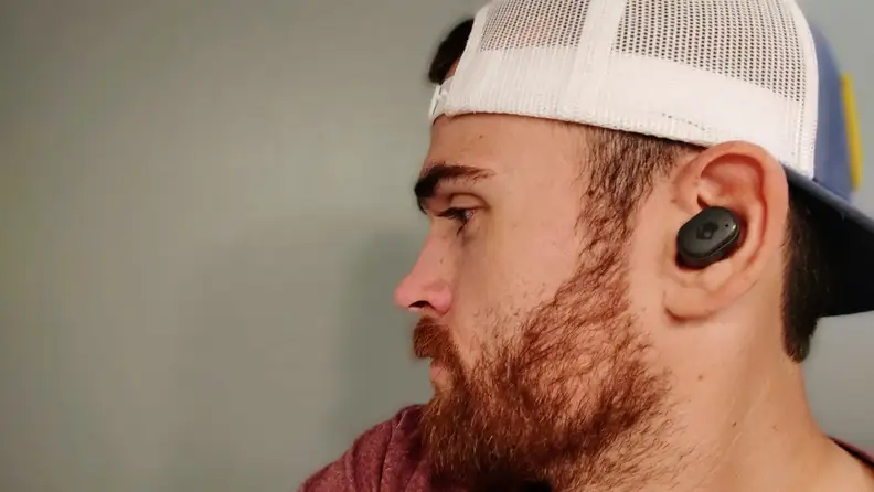 Skullcandy 's Grind Fuel all-black earbuds sit in the ear of a man in a baseball camp with a red beard.