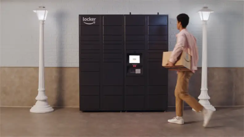 A person in a pink shirt and brown pants holding a box walks up to a black wall of lockers