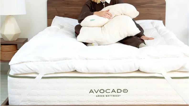 White Avocado Green Mattress topper on a bed with a woman sitting on top of it holding a pillow.