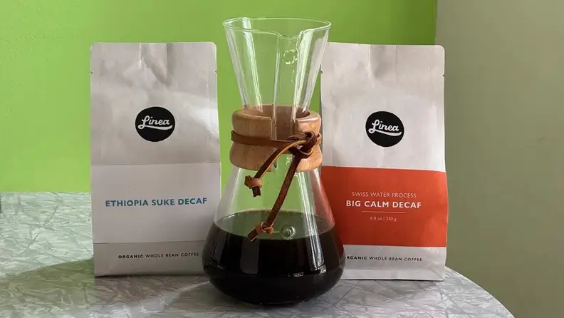 Two Linea decaf coffee bags with a glass carafe of brewed coffee
