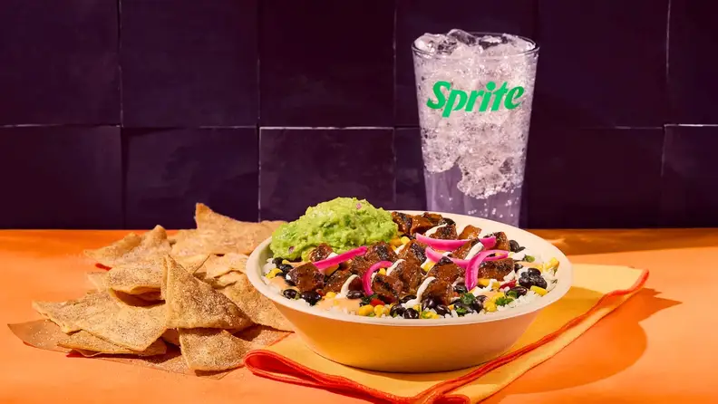 A loaded burrito bowl with steak, beans, corn, and guacamole, next to chips and a Sprite