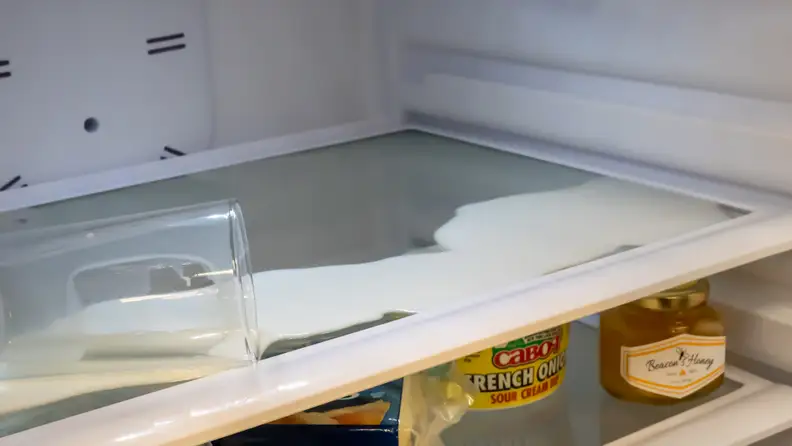 A close-up of the spill capture shelving in action. A glass of milk lays tipped over, spilling its contents out over the shelf. The milk is pooling at the edges of the shelf and not running over.