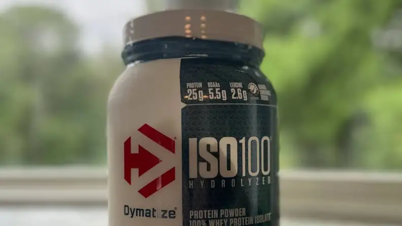 Close-up view of a bottle of Dymatize protein powder in front of a lush green background.