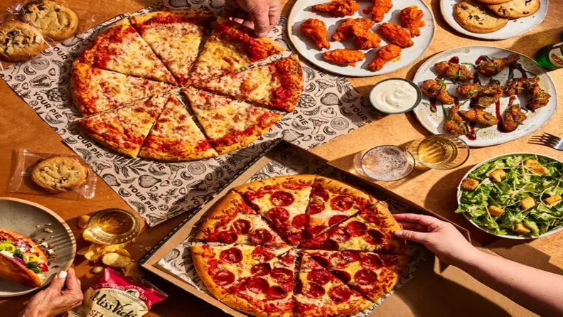 Pizzas, wings, salad, and cookies spread across a table with hands reaching in