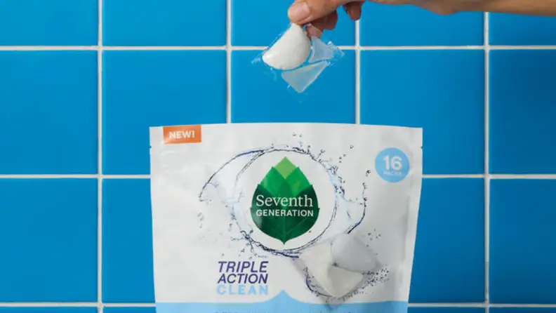 A dishwasher pack held above a bag of Seventh Generation dishwasher packs with a blue kitchen blacksplash