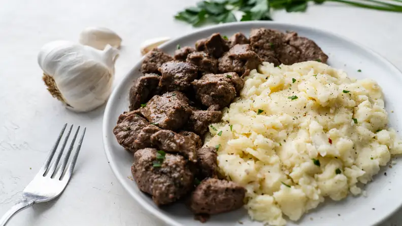 White plate with chopped beef and mashed potatoes next to a full clove of garlic