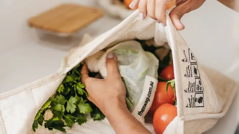 A person places vegetables inside of a canvas The Swag reusable produce bag.