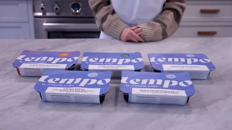 Five pre-packaged Tempo meals inside of individual containers on top of countertop surface in front of person with their hands folded together.