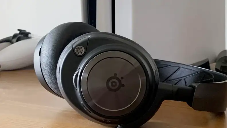 The Steelseries Arctis Nova Pro sit on a wooden surface in front of a PlayStation 5 and game controller.