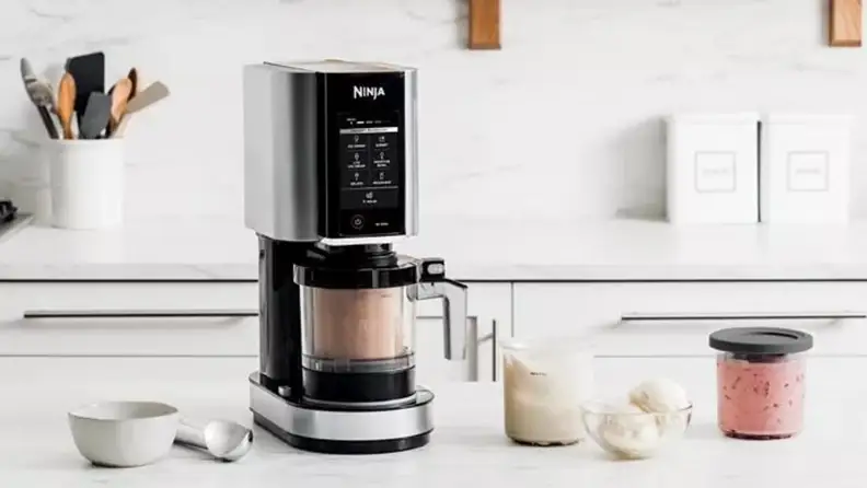 A ninja creami machine sits on a counter with ingredients bowls and an ice cream scoop