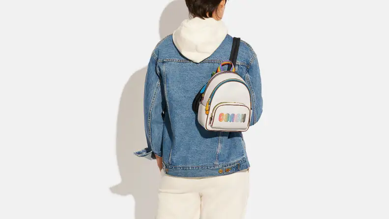 Person facing away wearing denim jacket, white pants, and white hoodie under jacket, and a white and black mini backpack that has "COACH" written across it in rainbow colors