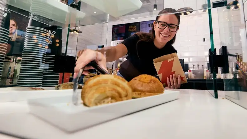 Smiling barista picking up a croissant with tongs behind a pastry case