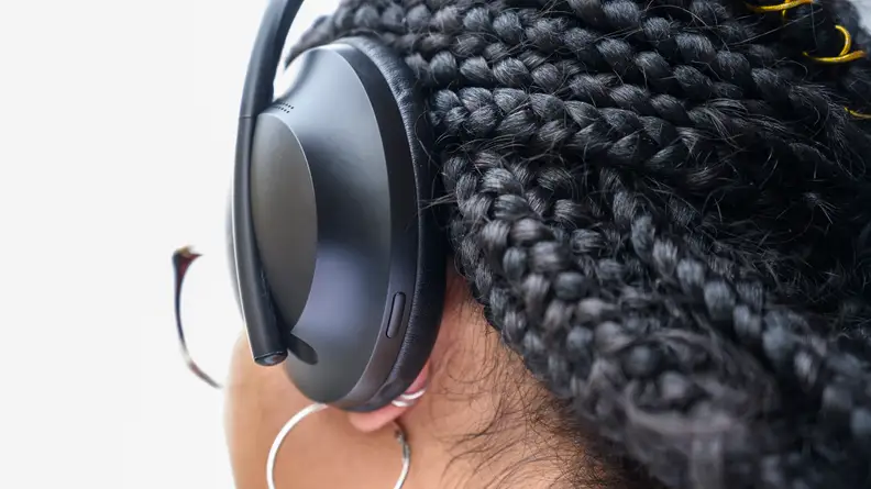 The back of a woman's head, wearing the Bose 700 wireless active noise-cancelling headphones. We can see a few small buttons along the  outside edge of the ear cups.