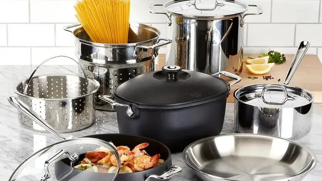 Assorted black and silver cookware set on kitchen countertop with raw pasta and cooked shrimp inside.