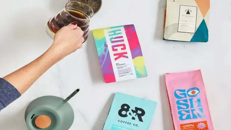 Hand pouring coffee next to assorted colorful coffee bags on a white surface