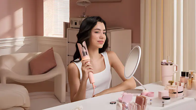 A woman using a pink automatic hair curler at a vanity mirror.