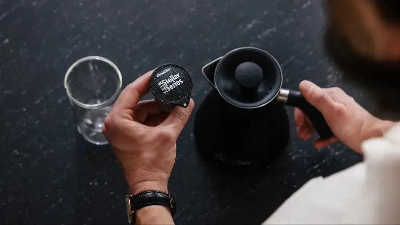 Hands holding a Cometeer Stellar Series coffee pod beside a black kettle and empty glass