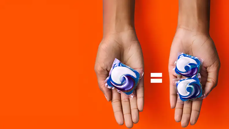 Two hands each hold laundry pods against an orange background, one hand holding a single pod and the other holding two smaller pods.