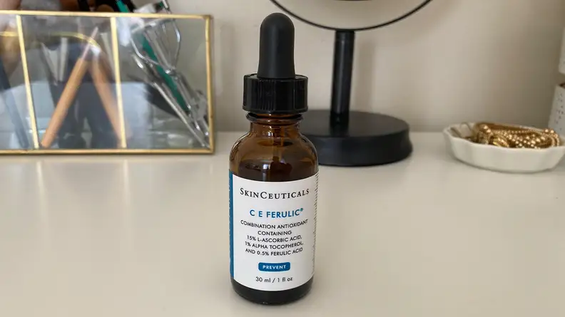 An amber glass bottle of skin serum with a white label standing on a white vanity desktop.