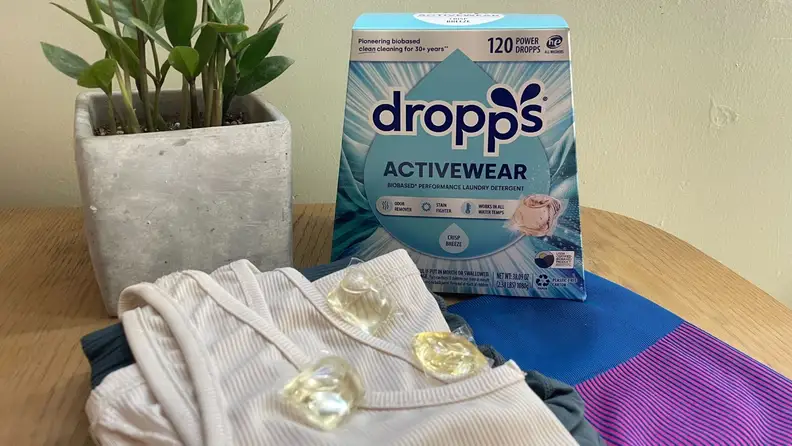 A box of Dropps detergent sits on a table with some clothes and a plant