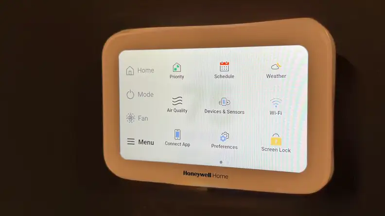 A Honeywell X8S smart thermostat on a maroon wall showing the home dashboard screen with various functions.