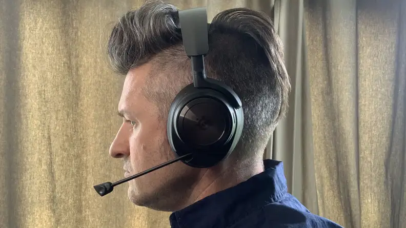 A Caucasian man in a dark blue shirt stands in front of green curtains while wearing the SteelSeries Arctis Nova Pro Wireless gaming headset with its microphone out.