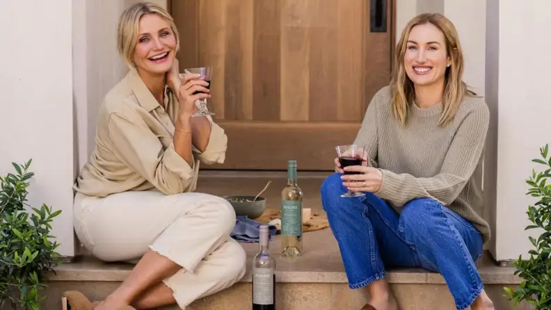 Cameron Diaz and Katherine Power posing on a stoop in front of a wood door and enjoying a glass of Avaline wine.