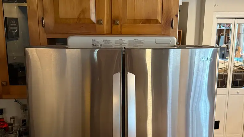 Top half of silver refrigerator shown with Bluetti FridgePower unit on top, in front of wooden cabinets.