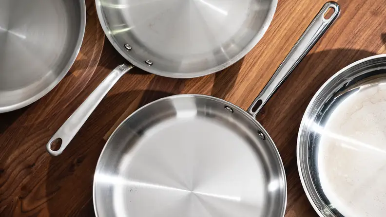 Four empty stainless steel skillets sit on a kitchen counter.