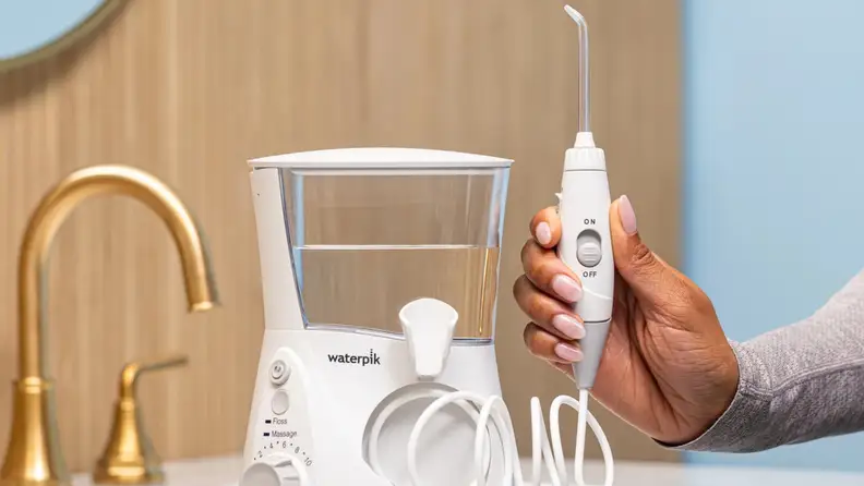 A white Waterpik Aquarius reservoir sits on a bathroom counter with a gold sink on the left and a hand holding the flossing mechanism on the right