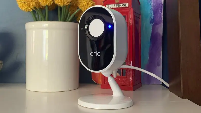 The Arlo Essential Indoor Security Camera sits on a white bookshelf with its camera privacy shield enabled.