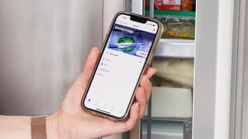 Person holding up smart phone to partially opened stainless steel refrigerator door with the Home Connect app on screen.
